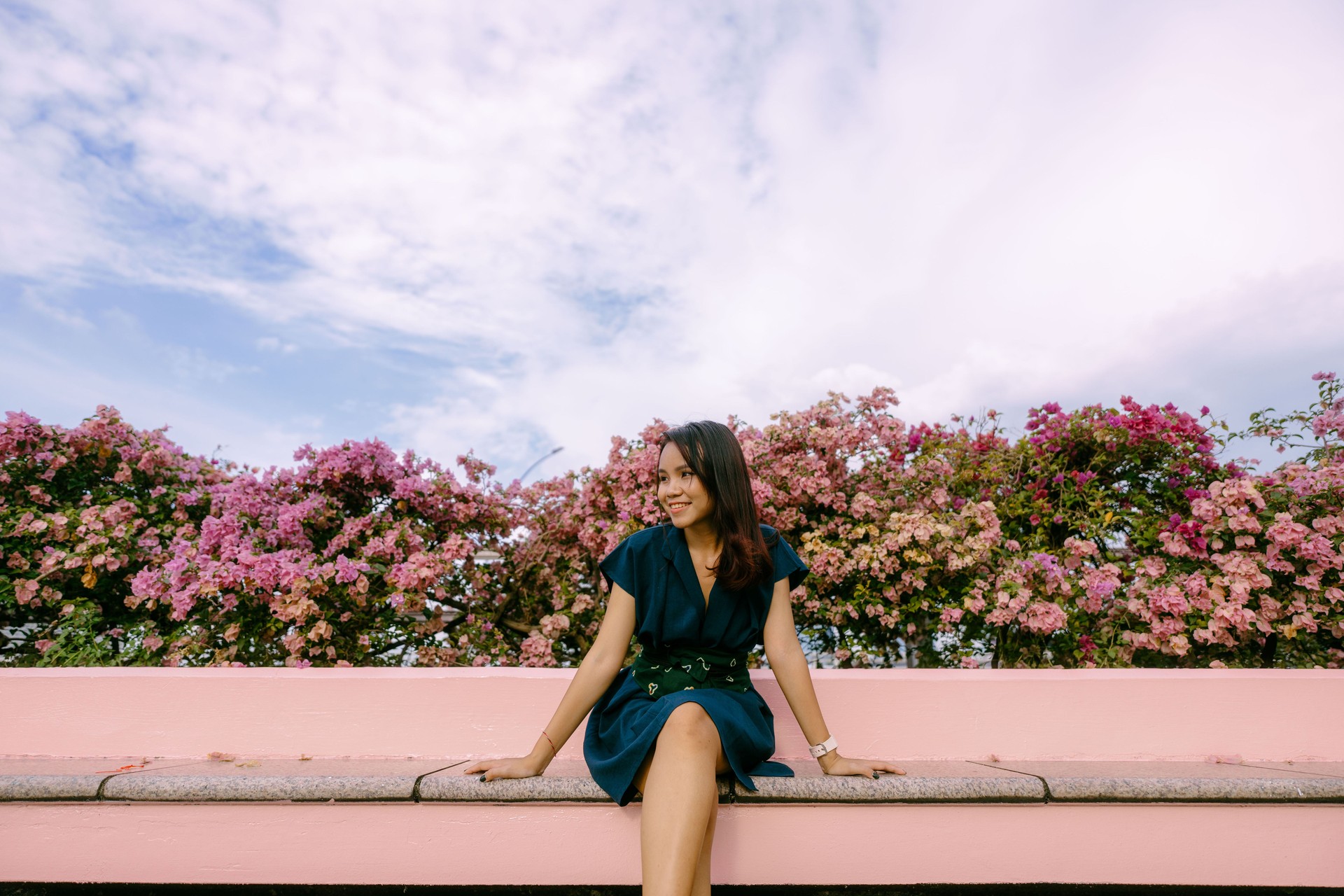 Asian Woman in Green Dress and Batik Obi Belt sitting Enjoying the Sun at sidewalk full of Pink Flowers